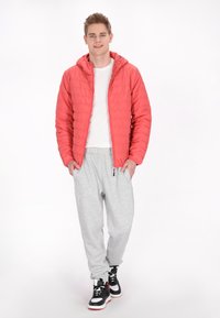 Rød quilted pufferjakke med hætte, hvid t-shirt, grå sweatpants og sorte sneakers med røde detaljer, stående foran en ensfarvet baggrund.