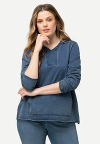 Modrá džínová mikina s v-výstřihem, dlouhými rukávy a přední kapsou. Má strukturovanou tkaninu a kontrastní švy podél lemu.