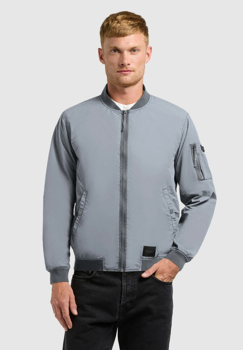 Homme aux cheveux courts portant une veste bomber grise zippée et un pantalon noir, se tenant devant un fond clair uni.