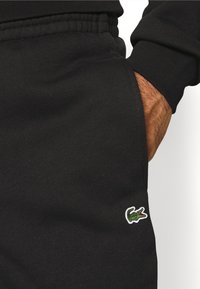 Main droite dans la poche d’un pantalon de survêtement noir avec taille élastique et logo crocodile vert brodé près de la poche.