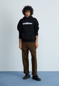 Sweatshirt preto com capuz com "the new originals" impresso na parte da frente, combinado com calças castanhas e sapatos pretos, em pé sobre um tapete azul.