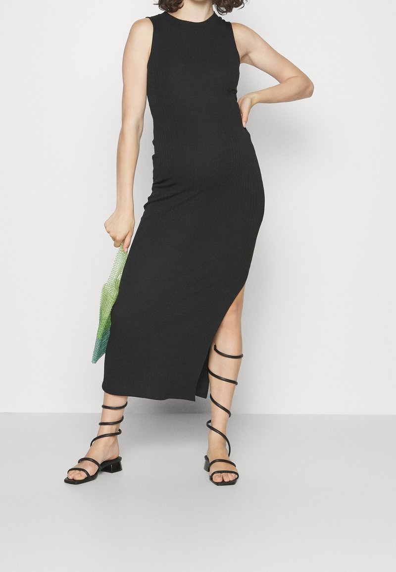 Femme portant une robe midi noire sans manches avec une fente latérale, des sandales noires à lacets, et tenant un petit sac à main dégradé vert.