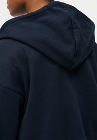 Φούτερ με κουκούλα σε ναυτικά μπλε, κατασκευασμένο από μαλακό, υφασμ ένο ύφασμα. Η κουκούλα είναι έντονη και οι ώμοι έχουν άνετη εφαρμογή.