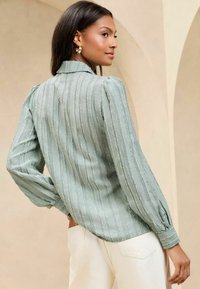 Blouse texturée vert clair avec des rayures verticales, manches longues bouffantes et un col. Associée à un pantalon crème.