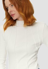 Haut à manches longues blanc à côtes avec col montant, présentant des détails de couture verticale et un design ajusté. Texture lisse et style minimaliste.