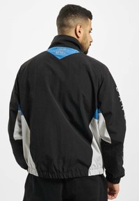 Black and white lightweight jacket with blue accents, standing collar, elastic hem, and sporty design. Features logo on upper back.