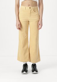 Pantaloni a gamba larga in corduroy giallo con una texture a rilievo, vita alta e tasche frontali. Abbinati a sneakers con motivo nero e grigio.