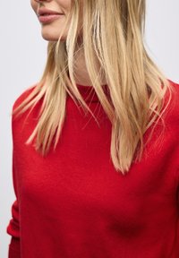 Maglione rosso lavorato a maglia con collo alto e maniche lunghe, caratterizzato da un tessuto testurizzato. I capelli biondi coprono parzialmente il maglione.