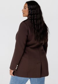 Bruine gestructureerde blazer met een getailleerde pasvorm, voorzien van een reverskraag, vier zilveren knoopaccenten op de mouw en details van een rugventilatie.