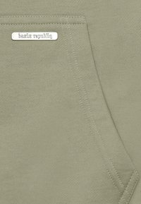 El tejido del sudadera verde claro presenta un bolsillo frontal con una etiqueta plateada grabada con "bazix republic". Textura suave y costuras cosidas visibles.