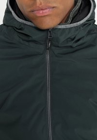 Chaqueta verde oscuro con cierre frontal, que presenta una capucha forrada en gris y acentos grises en contraste. Textura suave, diseño ligero.