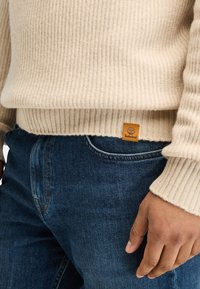 Maglione beige a coste con un'etichetta del logo Timberland visibile, indossato sopra jeans blu. Il maglione presenta un motivo a maglia testurizzato e polsini aderenti.