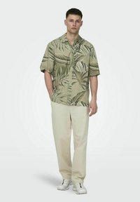 Kortlevenshirt in lichtgroen met een palmbladpatroon, gecombineerd met beige broek en witte sneakers. Casual outfit met een ontspannen fit.