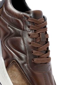 Zapatillas de cuero marrón con cordones marrón oscuro, que presentan paneles texturizados, un acabado liso y una suela de goma blanca. Detalles de costura acentuados visibles.