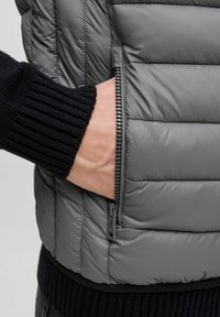 Pelēks izšūts veste ar rāvējslēdzēja sānu kabatu. Tuvplāns parāda roku, kas sniedzas iekšā kabatā, ar kontrastējošu melnu ribotu džemperi apakšā.