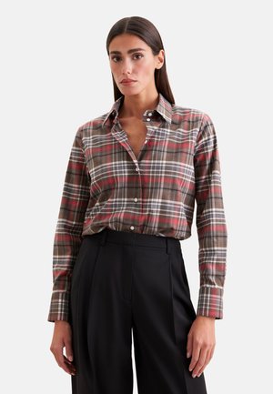 Femme portant une chemise à carreaux marron et rouge boutonnée, rentrée dans un pantalon noir taille haute à jambes larges, debout devant un fond uni.