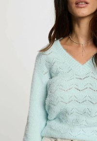 Maglione azzurro chiaro e soffice con scollo a V, caratterizzato da un motivo a onde e un design testurizzato. Una leggera lucentezza aggiunge profondità, indossato con una delicata collana d'argento.
