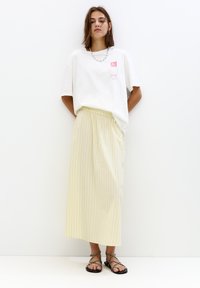 Witte oversized t-shirt met een rode graphic, gecombineerd met een lichtgele gestreepte midi rok en zwarte strapped sandalen.