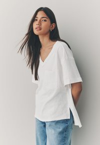 White V-neck T-shirt with short sleeves, featuring a front pocket and side slits, paired with blue denim jeans. Soft, textured fabric.