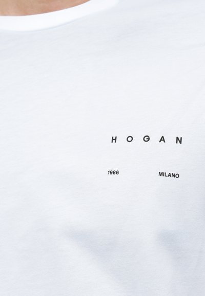 Λευκό βαμβακερό t-shirt με μικρό μαύρο λογότυπο που έχει το κείμενο "HOGAN", "1986" και "MILANO" τυπωμένα μπροστά. Ομαλή υφή.