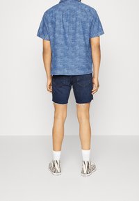 Blå kortärmad skjorta med vertikala ränder, i kombination med denimshorts. Personen bär vita strumpor och mönstrade sneakers.