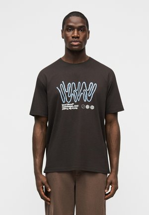 Homme portant un T-shirt noir avec des lignes abstraites bleues et un texte blanc "Wood Wood Copenhagen, 2002", debout devant un fond clair uni.