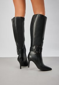 Bottes en cuir noires jusqu'au genou avec un bout pointu et un talon aiguille. Caractérisées par une large sangle en cuir avec un détail de boucle carrée.