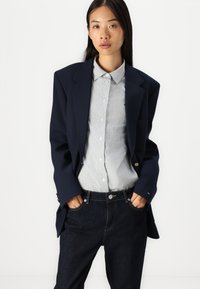 Blazer bleu marine avec des épaules structurées, associé à une chemise à rayures claires à boutons et un jean en denim foncé. Le blazer présente des accents de boutons dorés.
