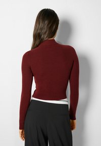 Suéter burdeos de cuello cisne acanalado con mangas largas, estilo ajustado y tejido texturizado, combinado con pantalones negros de cintura alta.
