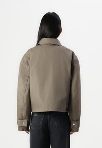 Giacca cropped khaki con colletto a punte, polsini a bottone e una texture liscia. Indossata sopra jeans in denim scuri. Design minimalista, vista da dietro.