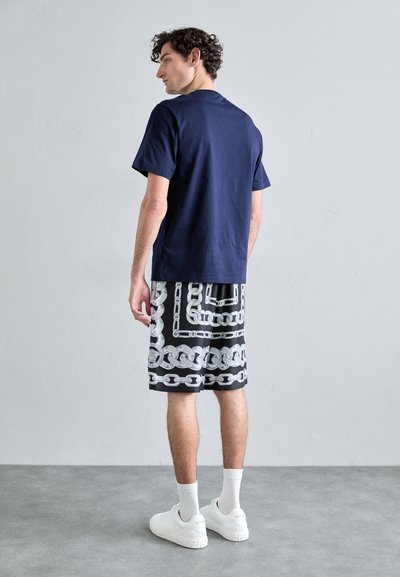 T-shirt en coton bleu marine associé à un short noir avec un motif de chaîne argenté ; des baskets et des chaussettes blanches complètent la tenue.