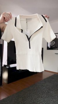 Vit kortärmad ribbad polo skjorta med svart kant på kragen och dragkedja, hålls uppe av händer i en modern vardagsrum.
