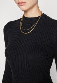 Guldpläterad kedja bärs över en svart ribbad stickad tröja med hög hals. Tröjan har en texturerad tyg och en åtsittande design.