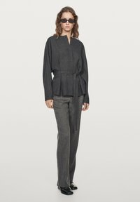 Giacca grigia in tessuto con cintura, dal taglio dritto e colletto alto, abbinata a pantaloni grigi su misura, indossata con scarpe nere.