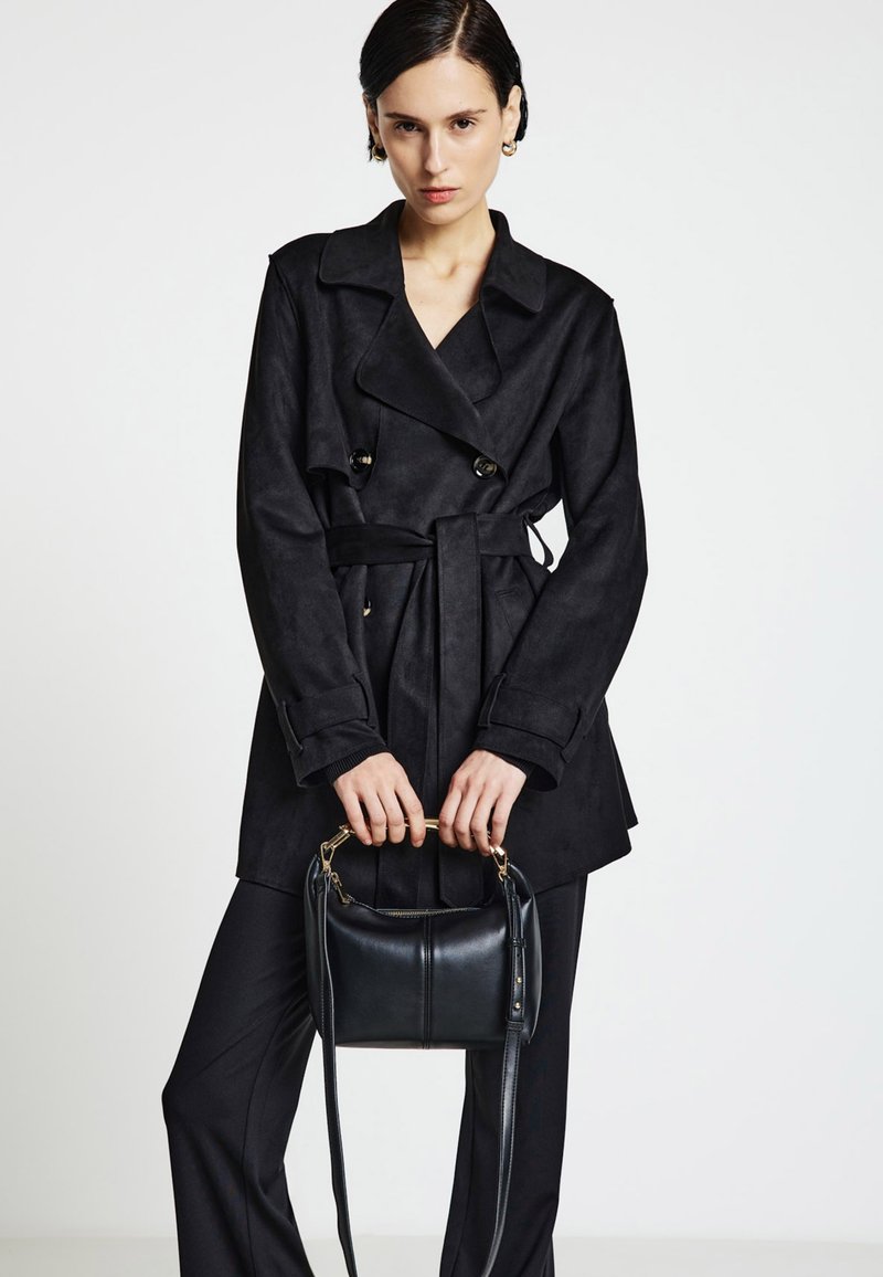 Trench coat nero a maniche lunghe in tessuto morbido con vita cinturata, abbinato a una piccola borsa in pelle nera con dettaglio manico dorato.