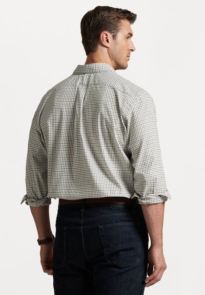 Mies, który ma podwinięte rękawy w kratkowanej koszuli i ciemnych dżinsach, stoi w pozycji skośnej, odwrócony plecami do aparatu na jednolitym tle.