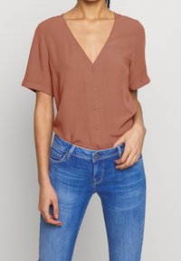 Blouse à manches courtes couleur rouille avec un col en V et des détails de boutons sur le devant, associée à un jean denim bleu ajusté. Texture de tissu lisse.