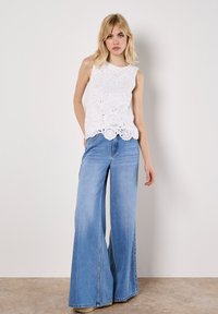 Top en dentelle blanche avec motifs floraux et ourlet en forme de scallop, associé à un jean bleu clair taille haute et à jambe large. Textures lisses, coupe décontractée.