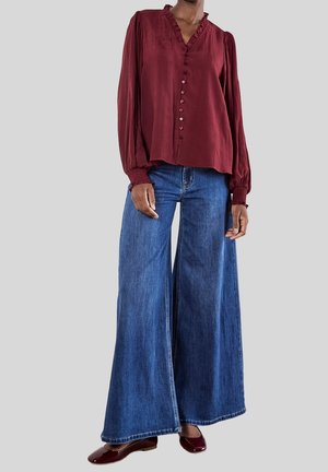 Vrouw draagt een bordeauxrode blouse met knopen, ruchekraag en wijde blauwe jeans, gecombineerd met bordeauxrode platte schoenen.