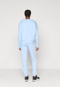 Osoba s vrkočmi stojí chrbtom, oblečená v svetlomodrom teplákovom oblečení s bielymi postrannými pruhmi a čiernymi športovými topánkami.