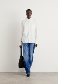 White button-up shirt, loose blue jeans, black bag. Smooth fabric, wide leg design, pointed black shoes, model stands against a plain background.
