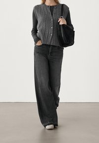 Graue Strickjacke mit Zopfmuster über einem dunkelgrauen Oberteil, kombiniert mit weit geschnittenen grauen Jeans und hellgrauen Turnschuhen; hält eine schwarze Schultertasche.