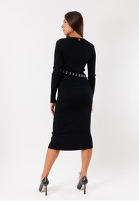 Vestito a maglia a coste nera con silhouette aderente, maniche lunghe e una cintura con accenti argentati in vita. Tacchi neri altezza caviglia.