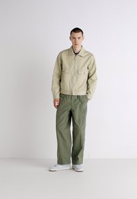Light beige zip-up jacket with two front pockets, paired with loose-fitting olive green trousers. Model wears white sneakers.