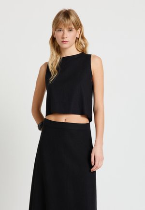 Giovane donna con capelli biondi che indossa un top nero senza maniche e una gonna nera a vita alta, in piedi davanti a uno sfondo chiaro e semplice.