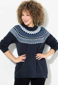 Kobieta z kręconymi włosami, ubrana w granatowy sweter z białym i niebieskim wzorem w okolicy szyi, z rękami na biodrach, uśmiechająca się na białym tle.