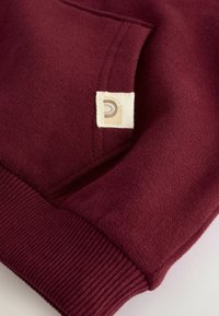 Pullover bordeaux con tasca a marsupio e polsini a coste. Un'etichetta quadrata con un logo a spirale è visibile sulla superficie del tessuto.