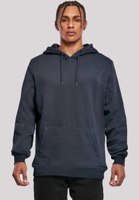 Young man with braided hair wearing a plain dark navy hoodie with front pocket and drawstrings, standing against a light gray background.
