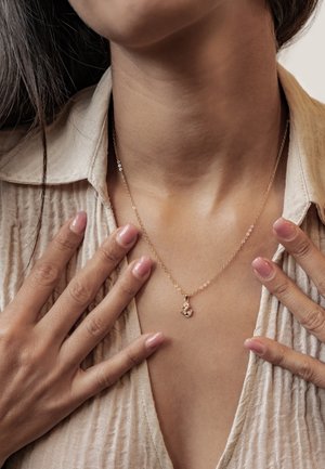 Close-up of a person wearing a delicate gold necklace with an anchor pendant, hands resting on an open light beige textured shirt.