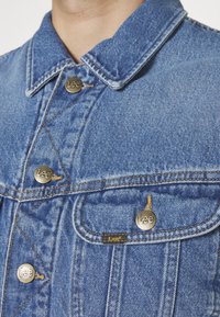 Denimjakke i mellemblå med spids krave. Har sølvknapper, kontrasterende syninger og et mærketag på lommen.